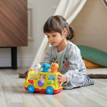 A young child sits on a light wood floor in a playroom, playing with a colorful toy bus. A cream teepee tent, a wooden cabinet, and a green cushion are visible in the background.