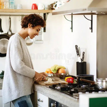 A woman with curly hair wearing a light gray knit sweater and blue jeans stands in a modern kitchen, preparing food at a wooden cutting board on a granite countertop.